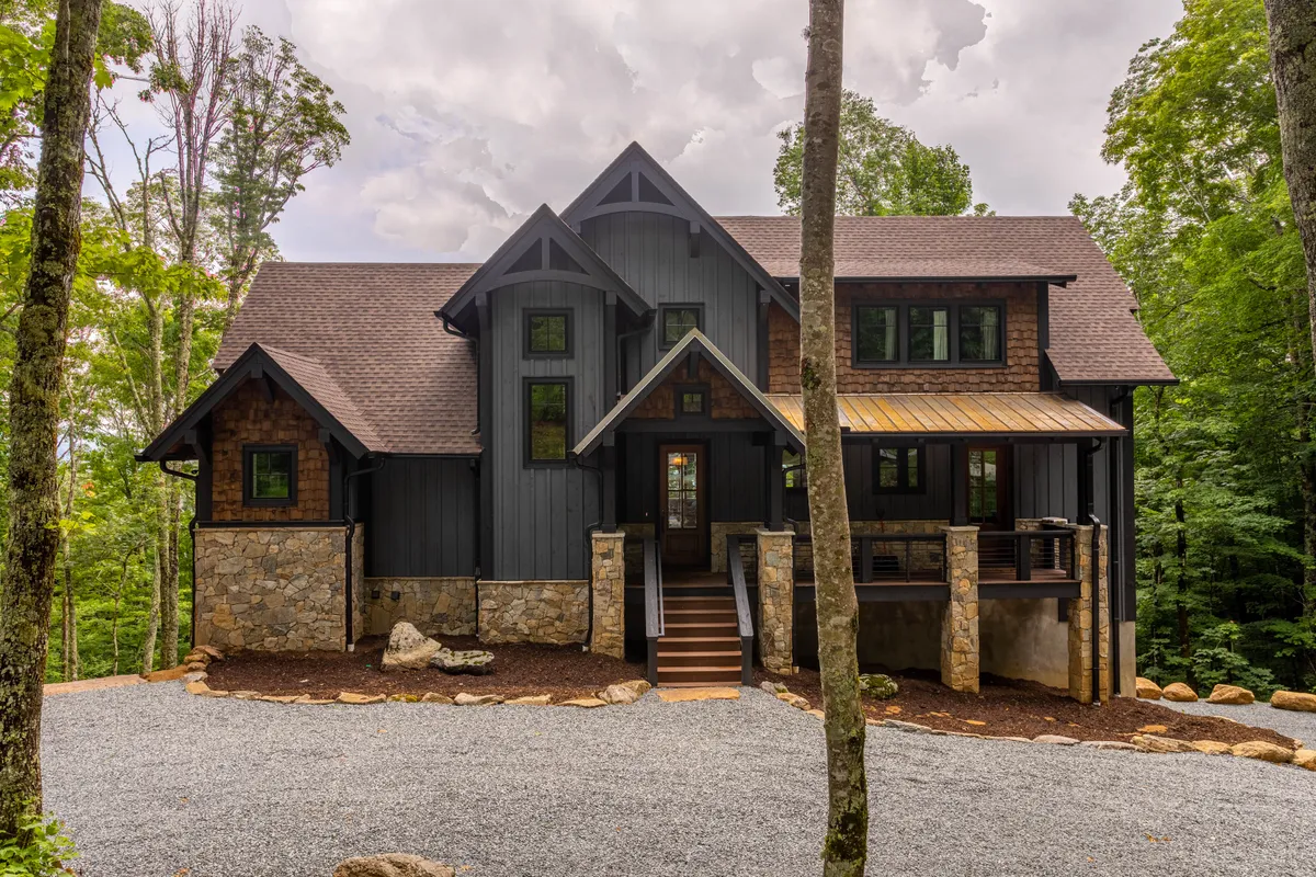 This image features the stone and wood facade of the cabin, with the unique roofline, front porch, and steps leading up to the entrance. The porch area is visible, set against a backdrop of peaceful woods, evoking a sense of welcome and connection to the natural surroundings.