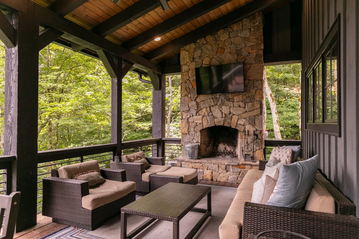 This exterior image presents a beautifully appointed outdoor living space with a large stone fireplace, comfortable seating, and a television. The scene is inviting, ideal for relaxing, socializing, or enjoying a quiet morning or evening outside.