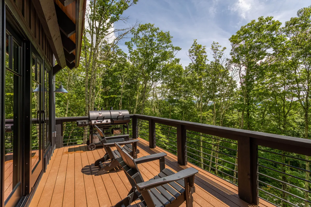 A wooden deck with deck chairs, a grill, and railing overlooks the surrounding trees. The deck has ample space for relaxing, barbecuing, or simply enjoying the fresh mountain air, with nature as the backdrop.
