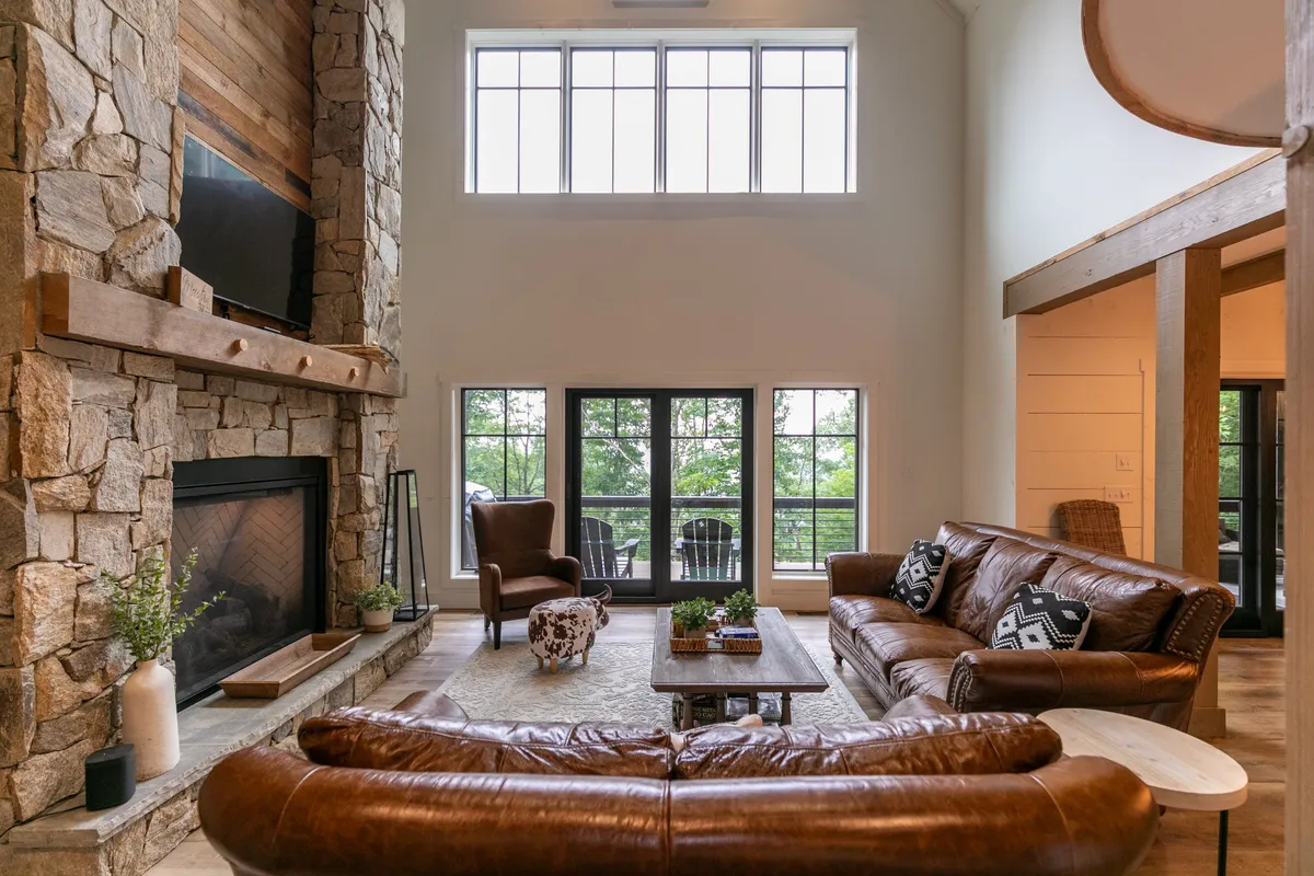 A beautifully styled open living room featuring a prominent stone fireplace, large windows with mountain views, leather sofas arranged for group seating, and a rustic wooden coffee table. Natural light pours in, highlighting the inviting atmosphere and spaciousness of the combined living area.