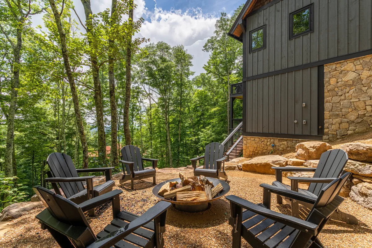 A cozy exterior fire pit area surrounded by Adirondack chairs and tall trees, set against the stone wall of the house. The fire pit setting is inviting, perfect for gathering at sundown for s’mores, storytelling, or simply relaxing beneath the stars.
