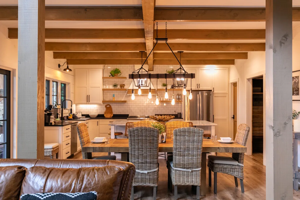 A view of the open dining area with a grand rustic wooden dining table, elegant chandelier overhead, and an open kitchen with visible granite island and wood beams. The setup encourages communal meals and conversation, with a stylish blend of modern and mountain elements.