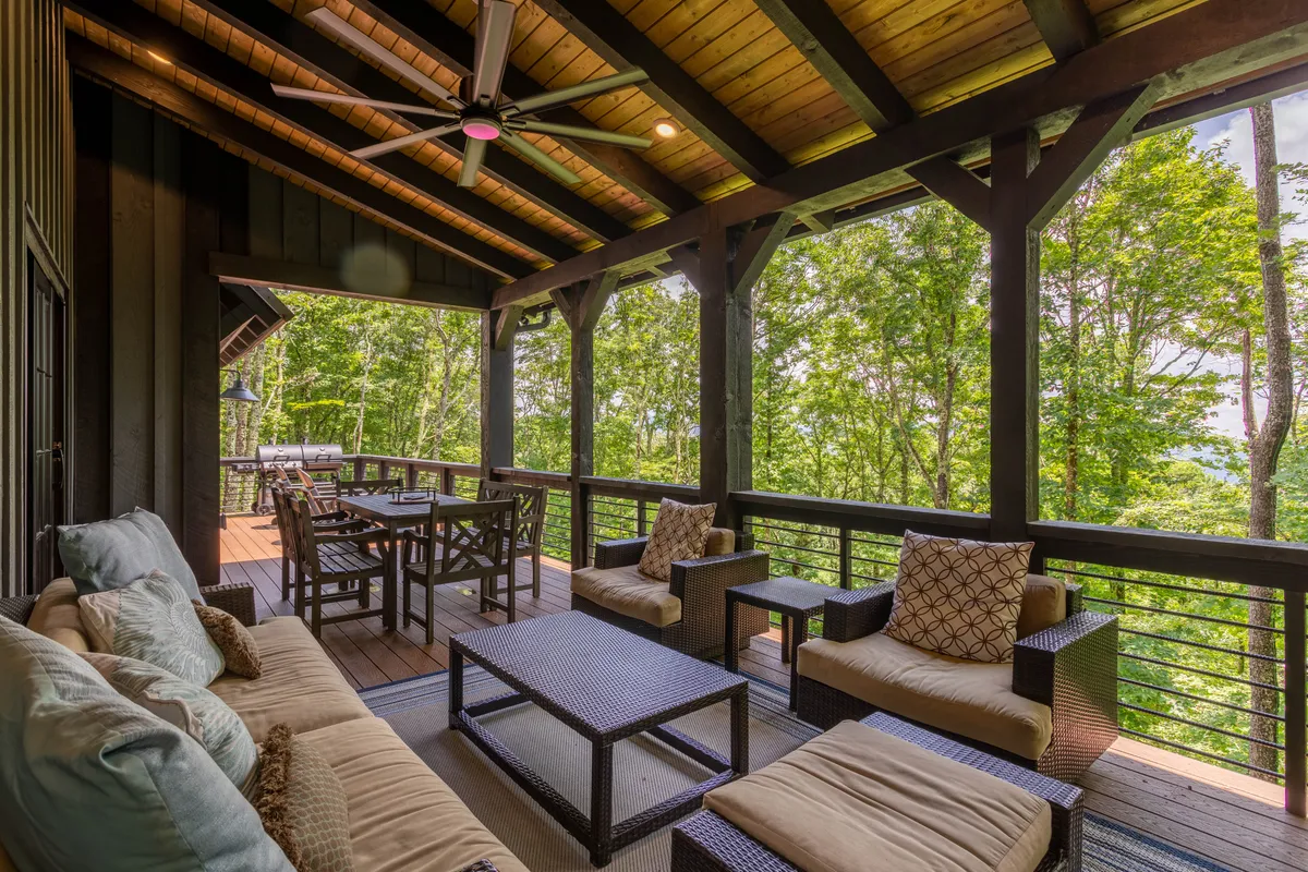 The image shows a comfortable outdoor seating area with views of lush trees, a dining table, and relaxed patio furnishings bathed in natural light. The deck is surrounded by woodland, inviting guests to dine or unwind outdoors with their pets.