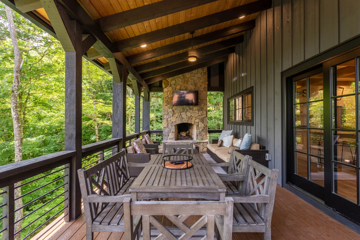 An expansive outdoor deck with a stone fireplace, comfortable seating area, dining table, and lush green surroundings. The setup is ideal for al fresco dining and relaxing by the fire, seamlessly extending the interior living space outdoors.