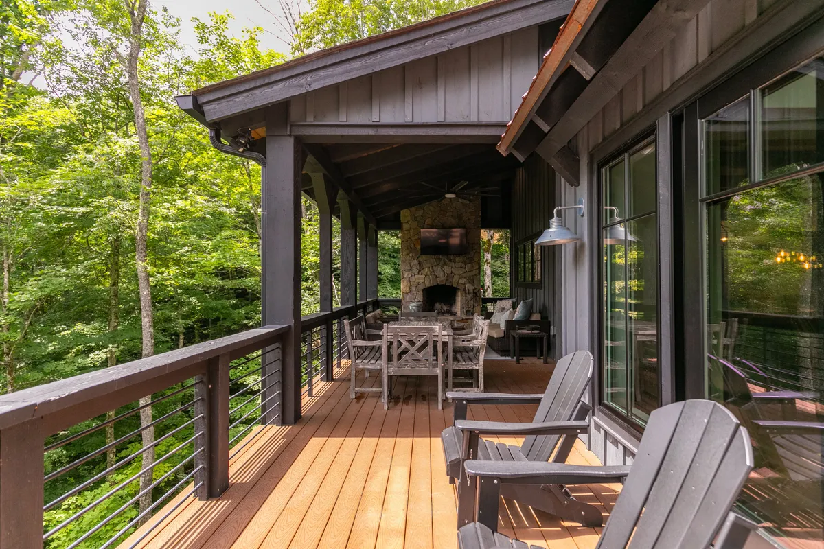 An inviting covered deck with a wooden outdoor dining table, surrounding trees, and a stone fireplace, creating a cozy spot for relaxation, yoga sessions, or intimate outdoor gatherings regardless of the weather.