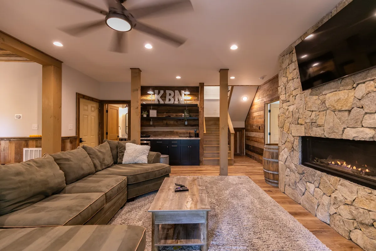 A spacious lower-level family room featuring a large sectional sofa, a stone fireplace, a kitchenette area, and inviting wooden accents. The scene is designed for relaxation, group gatherings, and entertainment, with cozy lighting that enhances the warm, welcoming atmosphere.