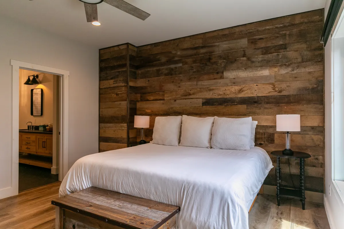 A serene bedroom featuring a cozy bed centered against a wooden wall, flanked by matching nightstands and a sunlit window. The space exudes calm and comfort, ideal for restful sleep or unwinding with a book.