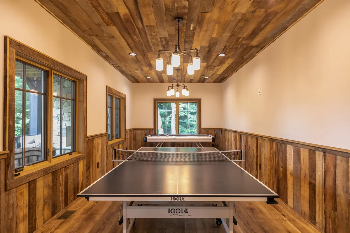 A vibrant, energetic interior game room with ping pong tables, a wooden ceiling, and large windows letting in natural light. The space is designed for fun and active family interaction, ideal for tournaments, board games, and creating lasting memories together.